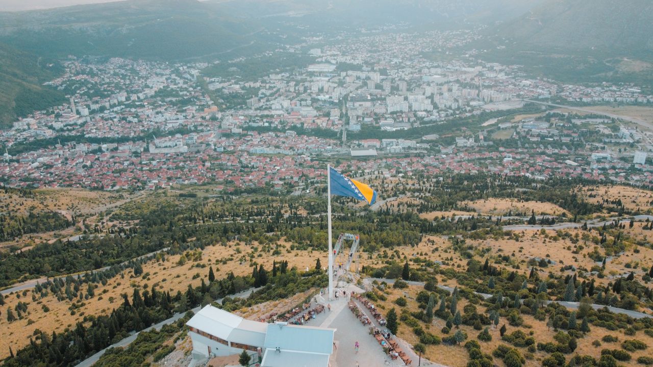 Scenic aerial shot of Mostar with Bosnia's flag prominently displayed, showcasing urban and natural landscapes.
